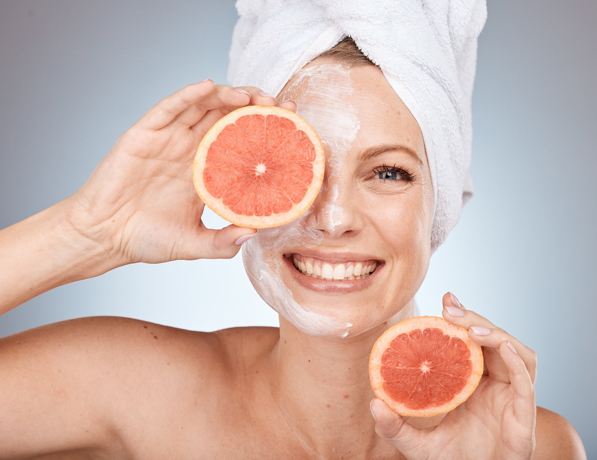 A woman smiling with face cream on while she holds slices of grapefruit in both of her hands, including one over her right eye.