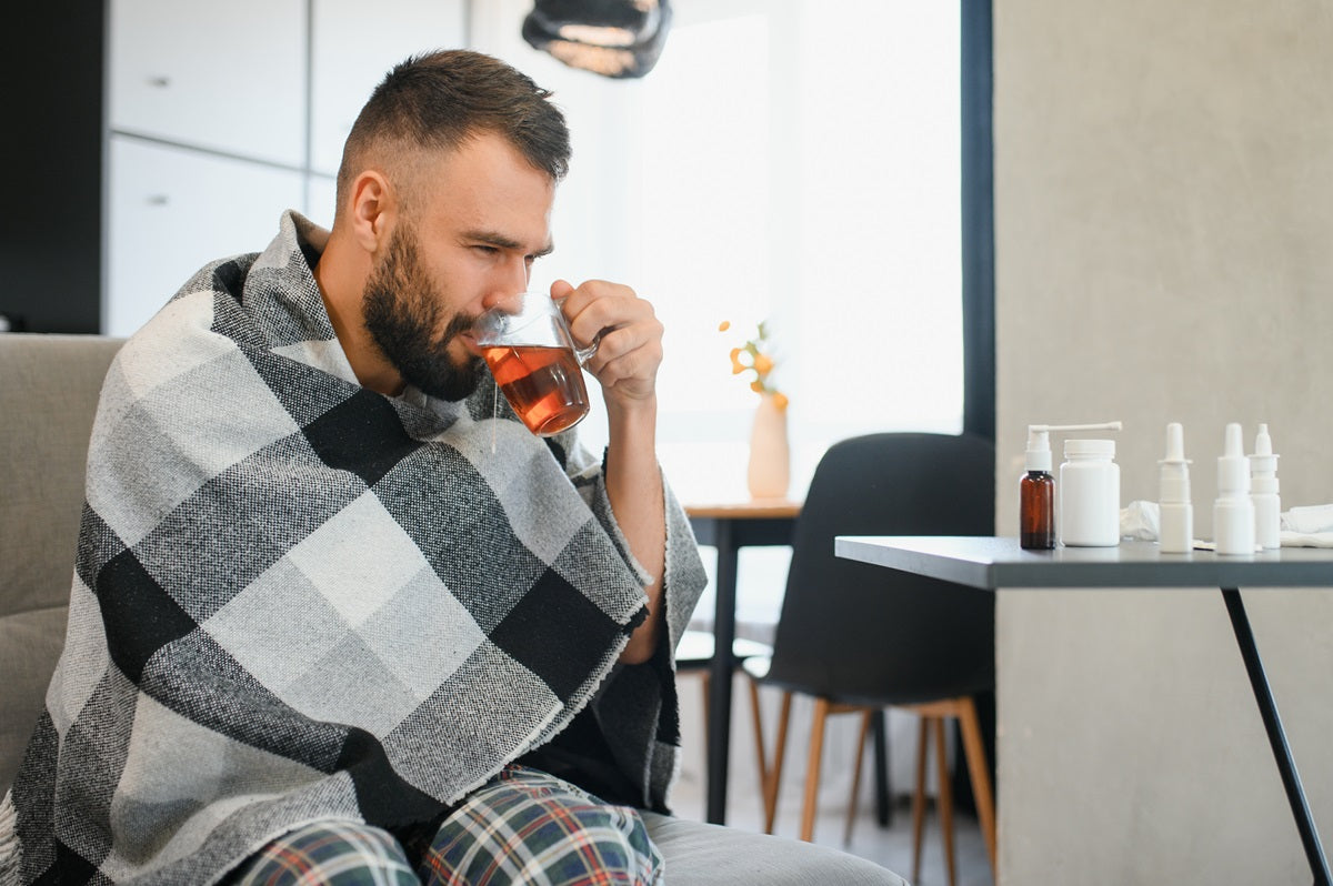 A sick man with a blanket over him sitting on the bed sipping tea
