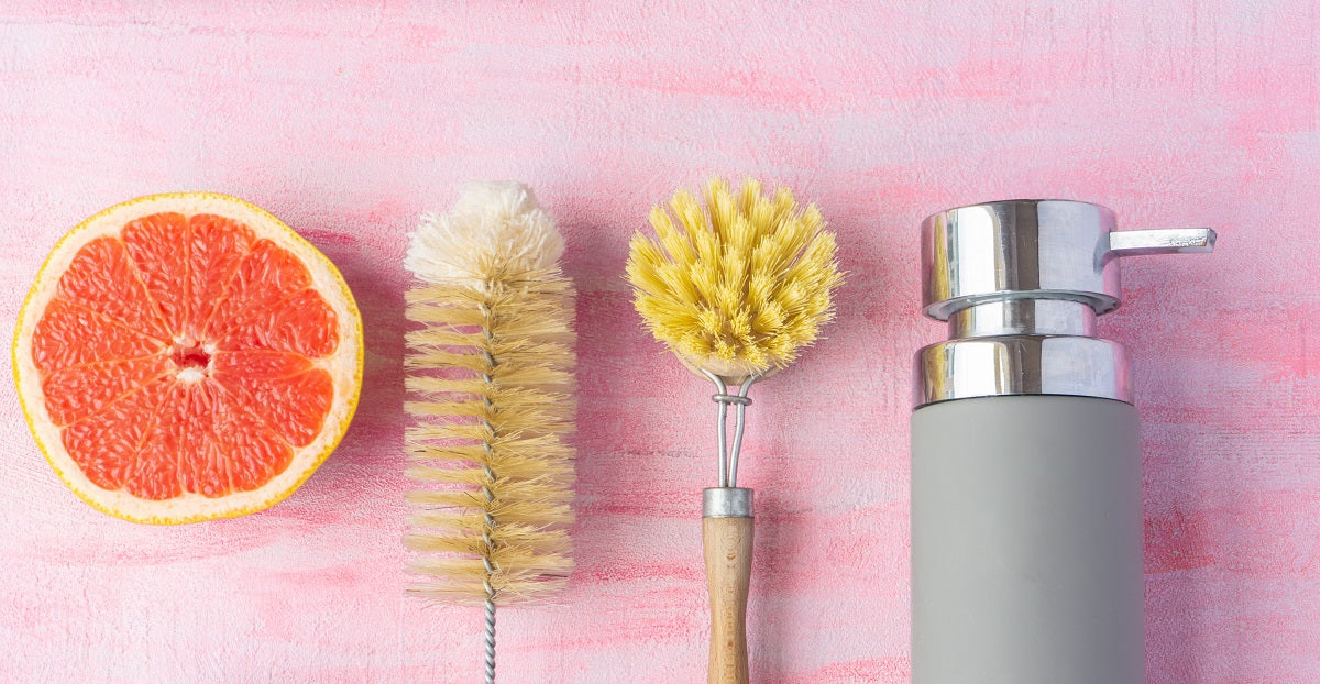 A grapefruit half rests next to a bottle brush, dish brush, and soap dispenser.