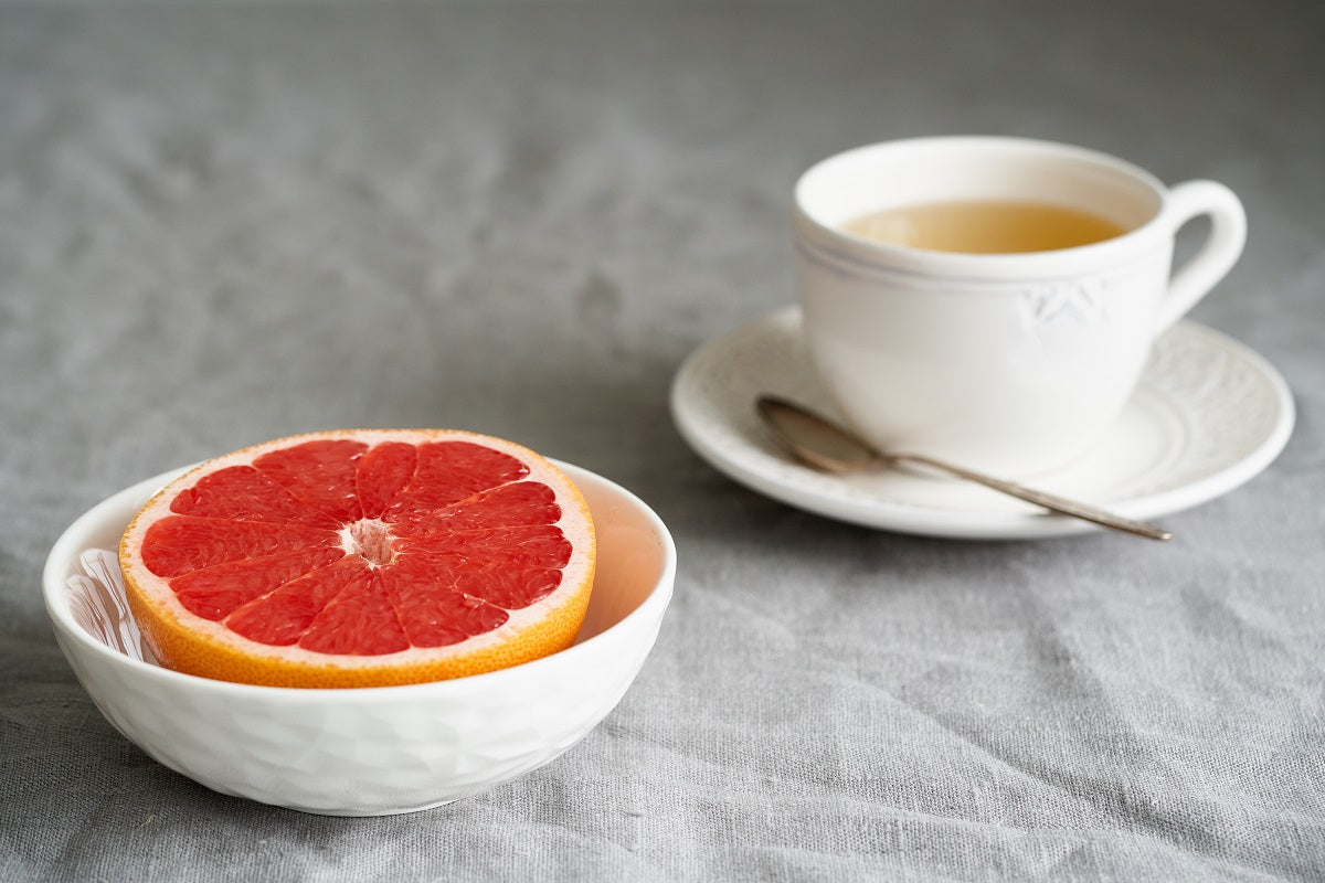 A grapefruit half sits in a bowl in front of a cup full of coffee on a saucer.