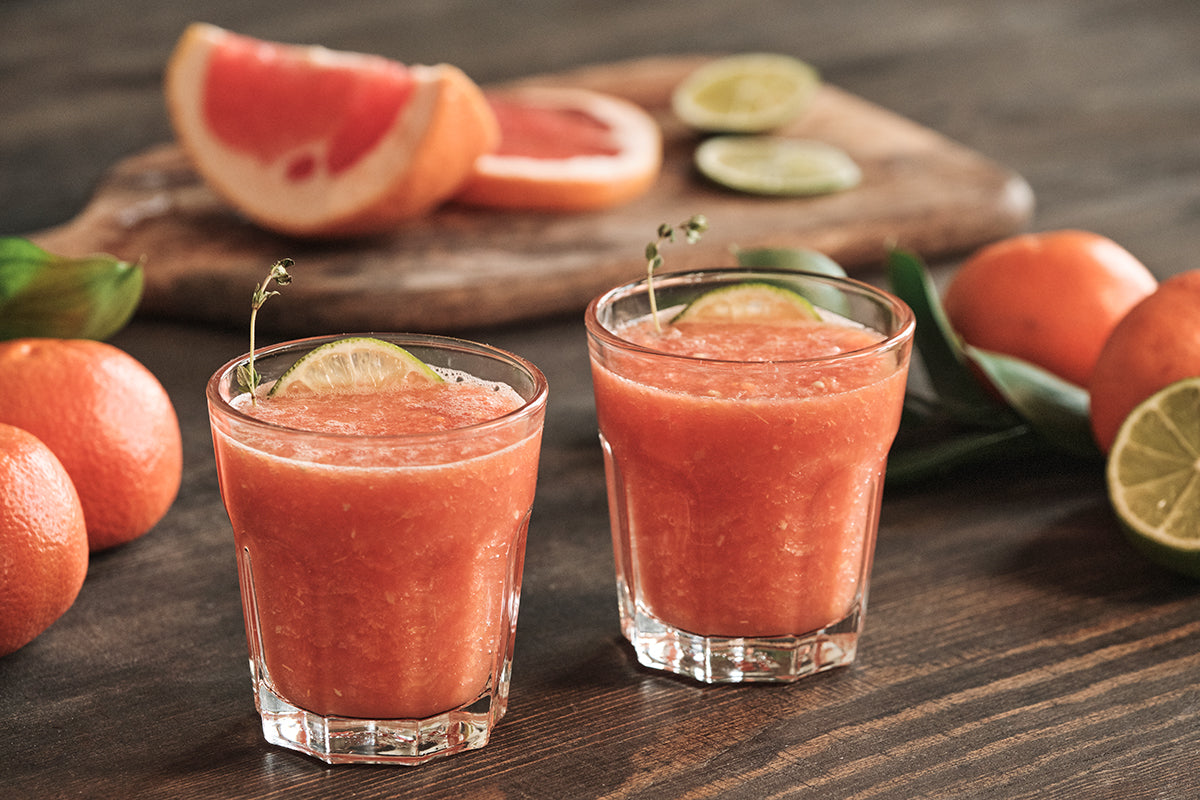 Red grapefruit smoothie in two glasses with whole grapefruits and grapefruit wedges scattered on the table